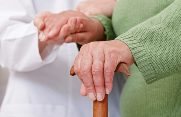 Physical therapist in a white coat holding the hand of a patient who is suffering with arthritis and is using a cane to stabilize themself